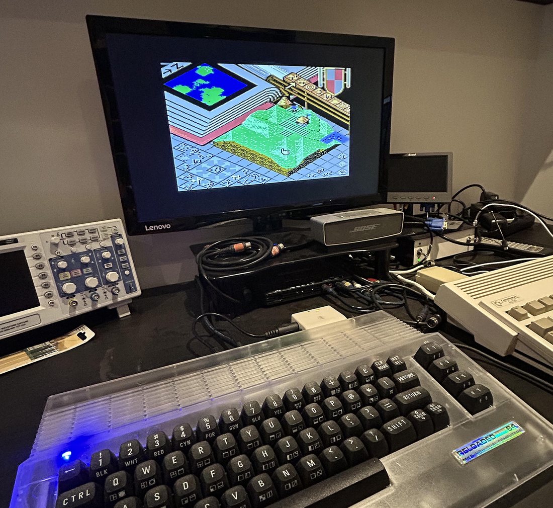 Populous running on a C64 Reloaded MK2 in Robert's workshop: a grass island on the monitor, the C64 keyboard in front, an oscilloscope beside it